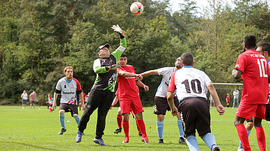 Der Fußball-Torhüter springt nach dem in der Luft befindlichen Fußball Er ist umringt von mehreren Fußballspielern auf einem Fußball-Rasenplatz.